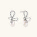Silver bow-shaped stud earrings with baguette crystals and dangling white freshwater pearls on cream background