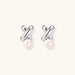 Rhodium-plated silver earrings with twisted X design and freshwater pearl drops on beige background