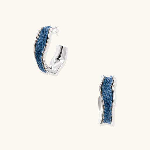 Silver huggie hoops with blue denim fabric inlay strip and polished white gold plated finish on cream background.